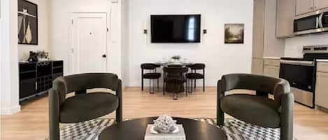 A modern living room with a round coffee table, two armchairs, a dining area with a table and chairs, a flat-screen TV mounted on the wall, and a kitchen area with cabinets and appliances.