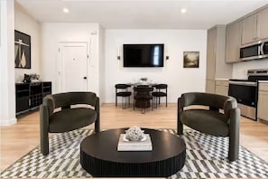 A modern living room with a round coffee table, two armchairs, a dining area with a table and chairs, a flat-screen TV mounted on the wall, and a kitchen area with cabinets and appliances. - King Bed-Unbeatable Location-Bishop Arts- Unit 3 (Dallas)