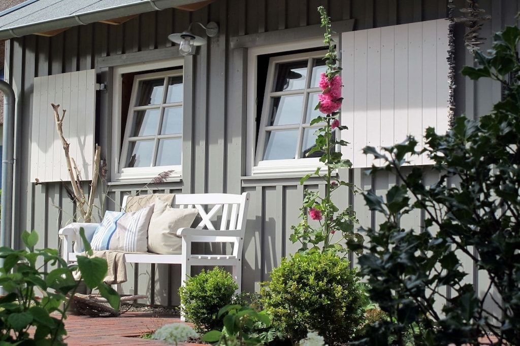 Une maison à l’extérieur blanc, un banc avec des coussins et, au premier plan, une plante à fleurs roses.