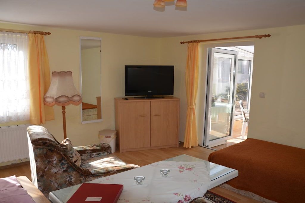 Una habitación con una televisión sobre un mueble de madera, un sofá, una mesa con mantel blanco y una ventana con cortinas.