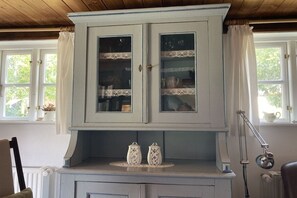 A room with a white cabinet with glass doors, a small table with two white ceramic containers, and a chair. - Holiday home in Born with swimming pool (Born)