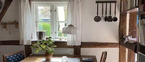 A wooden dining table with a white tablecloth, a potted plant, and a white vase. There is a window with white curtains and a view of greenery outside.
