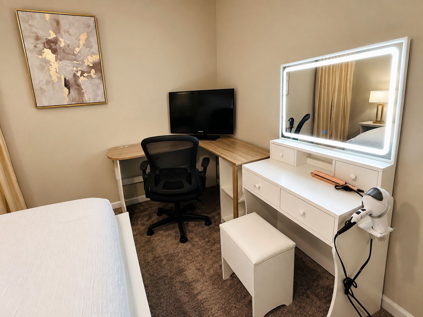 Habitación de hotel con cama, escritorio con silla, televisor, tocador con espejo y un taburete pequeño.