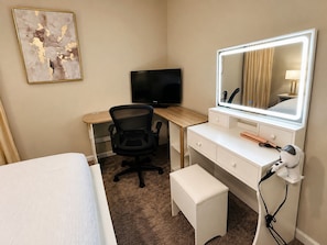 Quarto de hotel com uma cama, escrivaninha com cadeira, televisão, toucador com espelho e um banquinho pequeno.