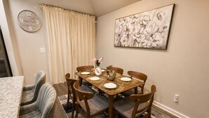 A dining area with a wooden table set for four, surrounded by chairs, a clock on the wall, and a painting above. - Modern Luxury 3BR Home in Cullman (Cullman)