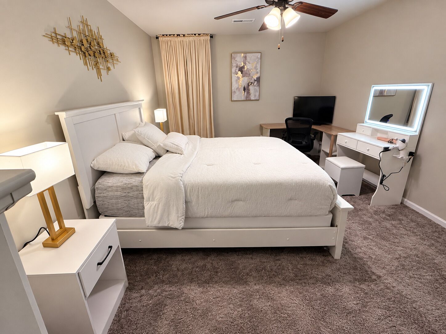 Un dormitorio con cama, mesitas de noche, escritorio, silla, ventilador de techo, televisor y espejo.