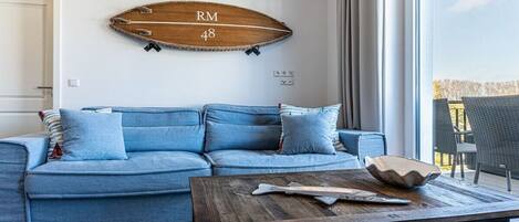 Un salon avec un canapé bleu, une table basse en bois et un élément de décoration murale en forme de planche de surf.