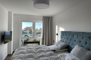 A bedroom with a bed, a television mounted on the wall, a window with curtains, and a view of the outside. - Holiday apartment with 1 bedroom (Kühlungsborn)