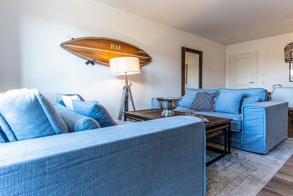 Ein modernes Wohnzimmer mit einem blauen Sofagarnitur, einem an der Wand befestigten Holzregal mit 'RM' und einer Lampe sowie einem großen Spiegel.
