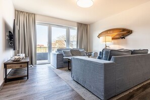 Ein modernes Wohnzimmer mit einem grauen Sofa, einem kleinen Tisch und einem Fernseher. Eine Glastür öffnet sich zu einem Außenbereich.