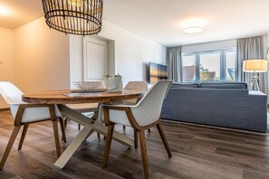 A modern living room with a wooden dining table, white cushioned chairs, a grey sofa, and a large window with curtains. - Holiday apartment with 1 bedroom (Kühlungsborn)