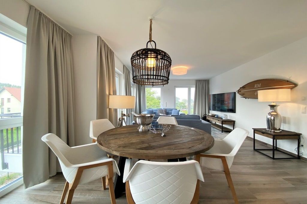 A modern living room with a round wooden table, white chairs, a hanging light fixture, and a TV mounted on the wall.