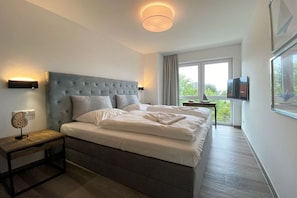 A modern bedroom with a tufted headboard, a bedside table with a lamp, a flat-screen TV, and a large window with curtains. - Holiday apartment with 1 bedroom (Kühlungsborn)