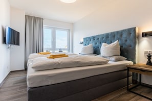 Habitación de hotel moderna con una cama grande, un televisor de pantalla plana montado en la pared, una ventana con cortinas y una mesita de noche con lámpara.