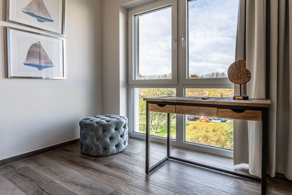 Een kamer met een groot raam, een bureau, een ottoman met gesponnen bekleding en twee ingelijste foto’s aan de muur.