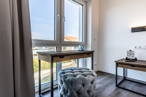 Ein Zimmer mit großem Fenster, einem Holztisch, einem samtenen Hocker und einem kleinen Schreibtisch mit Lampe.