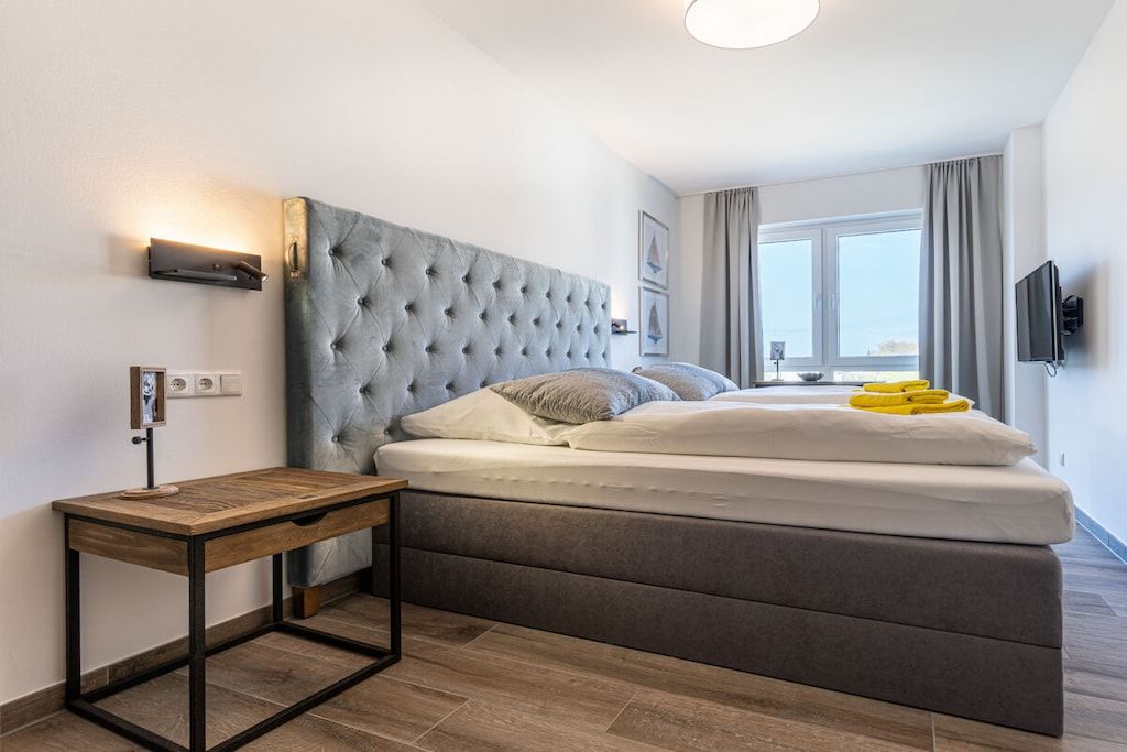 Ein Schlafzimmer mit einer gepolsterten Kopfstütze, einem Nachttisch aus Holz, einem Bett mit weißer Bettwäsche und einem Fenster mit Vorhängen.