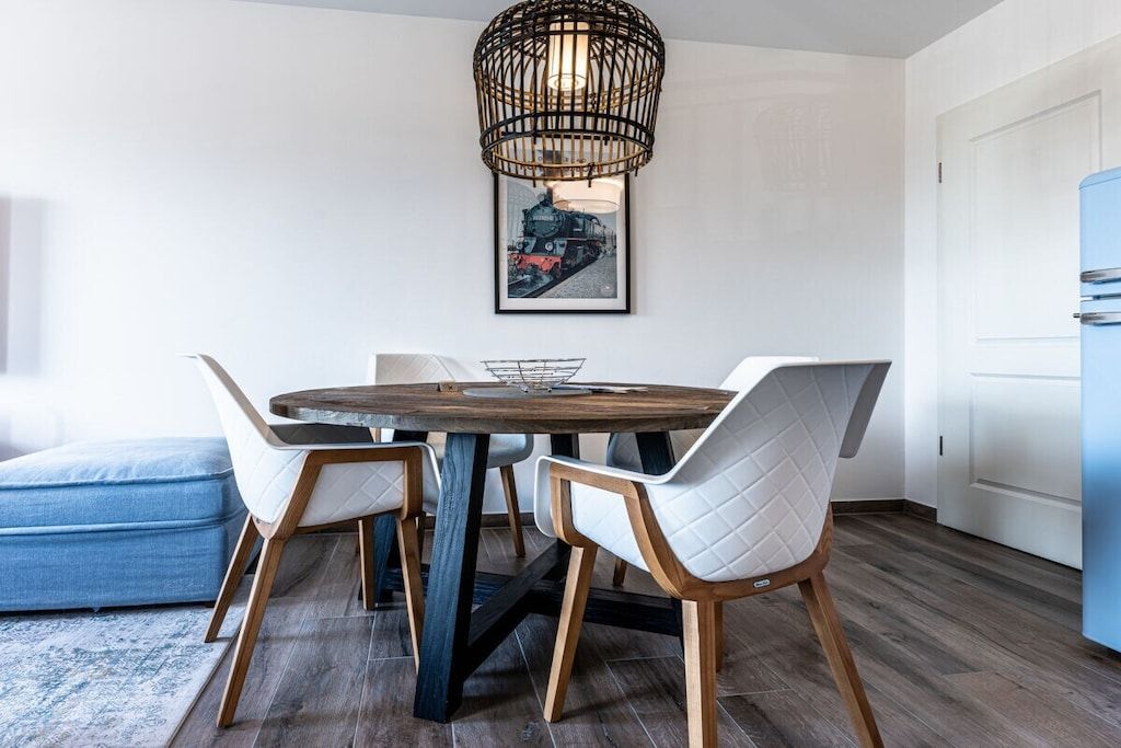 Un comedor con una mesa redonda de madera, sillas blancas con acolchado, una lámpara colgante de metal negro y un cuadro en la pared.