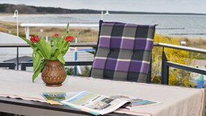 Ett bord med en vas med röda blommor och en tidning, en rutig kudde och utsikt över havet.