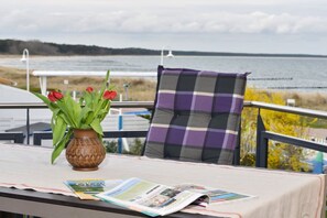 Een tafel met een vaas rode bloemen en een tijdschrift, een geruit kussen, en uitzicht op zee.