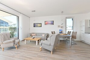 Ein modernes Wohnzimmer mit einer Couch, einem Esstisch und Stühlen. An der Wand hängen gerahmte Bilder und ein großes Fenster lässt viel Tageslicht herein.