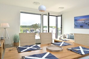 A dining area with a wooden table, wicker chairs, and a view of the outdoors through large windows. - Holiday apartment in Glowe with swimming pool (Glowe)