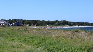 En strand med en modern byggnad i bakgrunden, en klar himmel och en grönskande förgrund.