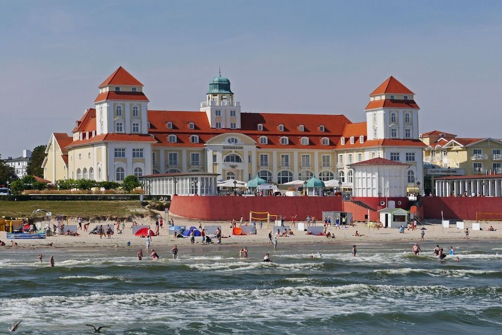Een groot gebouw met een rood dak en meerdere torens, grenzend aan een strand met mensen en parasols.