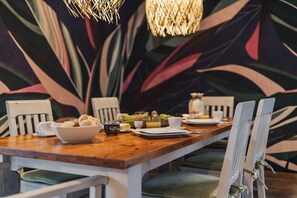 A dining table with a wooden top, white chairs, and a tropical leaf-patterned wall in the background. - Holiday home in Glowe with swimming pool (Glowe)