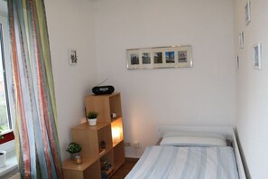 Ein Schlafzimmer mit einem Bett, einem Regal mit einem Radio, einem Fenster mit Vorhängen und einem an der Wand befestigten Bilderrahmen.