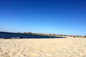 Une plage de sable fin, un ciel bleu dégagé, une mer calme et quelques bâtiments visibles au loin.