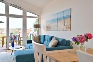 A modern living room with a blue sofa, a dining table, and a large window offering a view of the outdoors. - Holiday apartment in Glowe with swimming pool (Glowe)