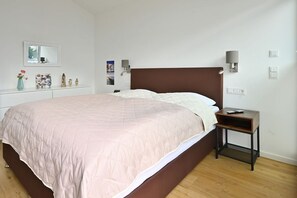 Un lit bien fait, avec une tête de lit, une table de chevet avec une lampe et une petite étagère ornée d’objets décoratifs.