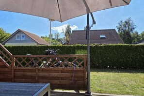 Eine Holzterrasse mit Metallgeländer, einem großen Sonnenschirm und freiem Blick auf ein Haus mit Ziegeldach im Hintergrund.