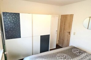Ein Schlafzimmer mit einem Bett, einem Schrank mit geometrischen und schlichten Paneelen, einer Holztür und einem Spiegel an der Wand.