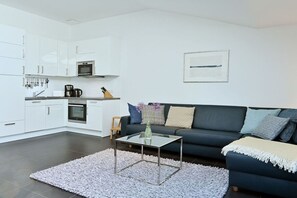 A modern living space with a kitchenette, a dark-colored sofa, and a glass coffee table. - Holiday apartment in Juliusruh with Wi-Fi (Juliusruh)