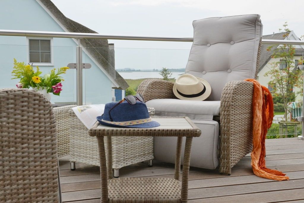 A wicker lounge chair with a cushion, a small table with a hat, and a view of a body of water.