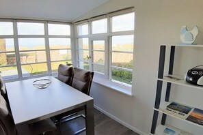 A corner room with a dining table, chairs, a white shelf with books, and large windows offering a view of the outdoors. - Jette beach bungalow (Glowe)