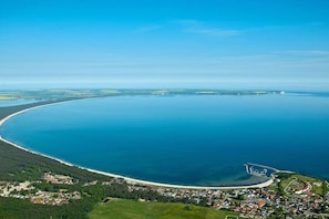 An aerial view of a coastal area with a large body of water, a curved coastline, and a small settlement with houses and greenery. - Jette beach bungalow (Glowe)
