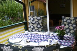 Ein runder Tisch mit kariertem Tischläufer, umgeben von Stühlen mit floralem Muster, und ein Blumentopf mit rosa Blüten.