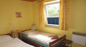 Ein kleines Zimmer mit einem Einzelbett, einem Fenster, einem Nachttisch, einem Stuhl und einem Teddybären.