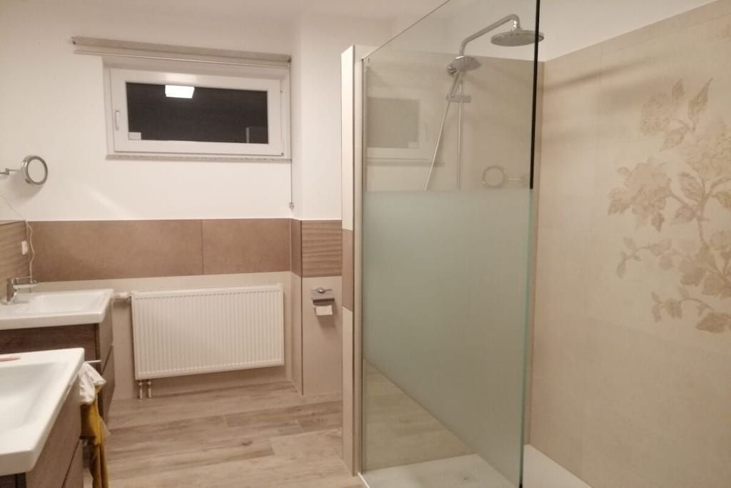 A bathroom with a glass shower enclosure, a white sink, and a window above the mirror.