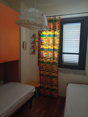 Een slaapkamer met twee bedden, een raam met zonwering, een kleurrijk gordijn en een hanglamp.