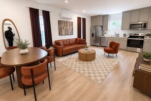 A modern living room with a brown leather sofa, a wooden dining table with orange chairs, a kitchen with stainless steel appliances, and a checkered rug.