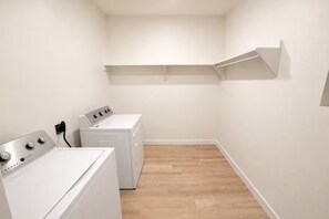 Un lavadero con dos lavadoras blancas y una secadora, piso de madera y pared blanca con estante.