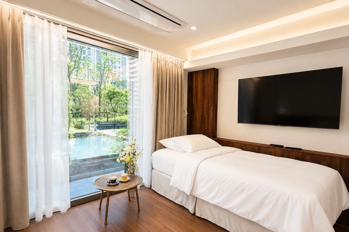 A hotel room with a bed, a TV, a small table with a cup, and a view of a pool and greenery outside.