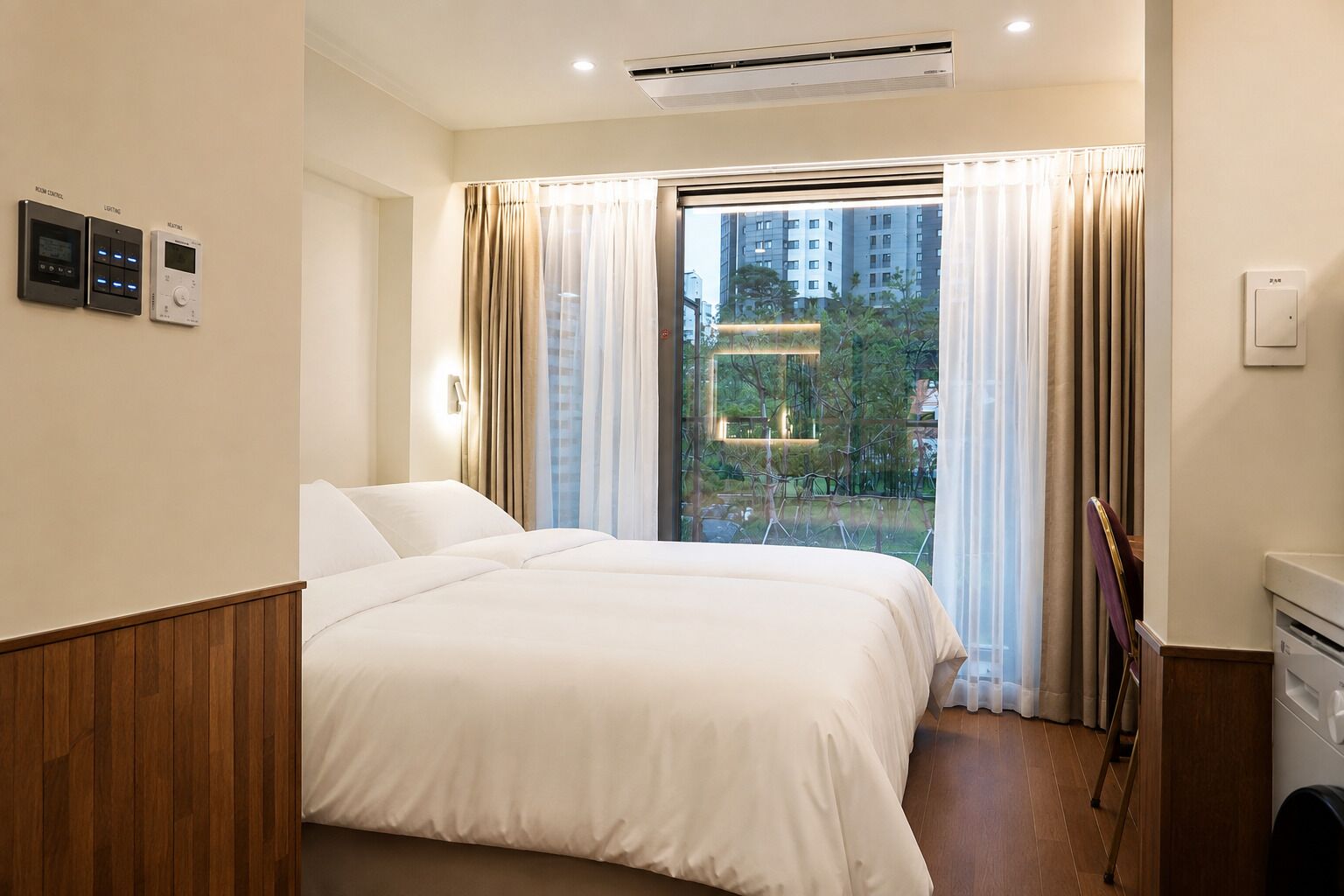 A hotel room with a bed, a window with curtains, an air conditioning unit, and a control panel on the wall.