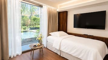 Quarto de hotel com cama, televisão, uma pequena mesa com uma chávena e vista para uma piscina e vegetação lá fora.
