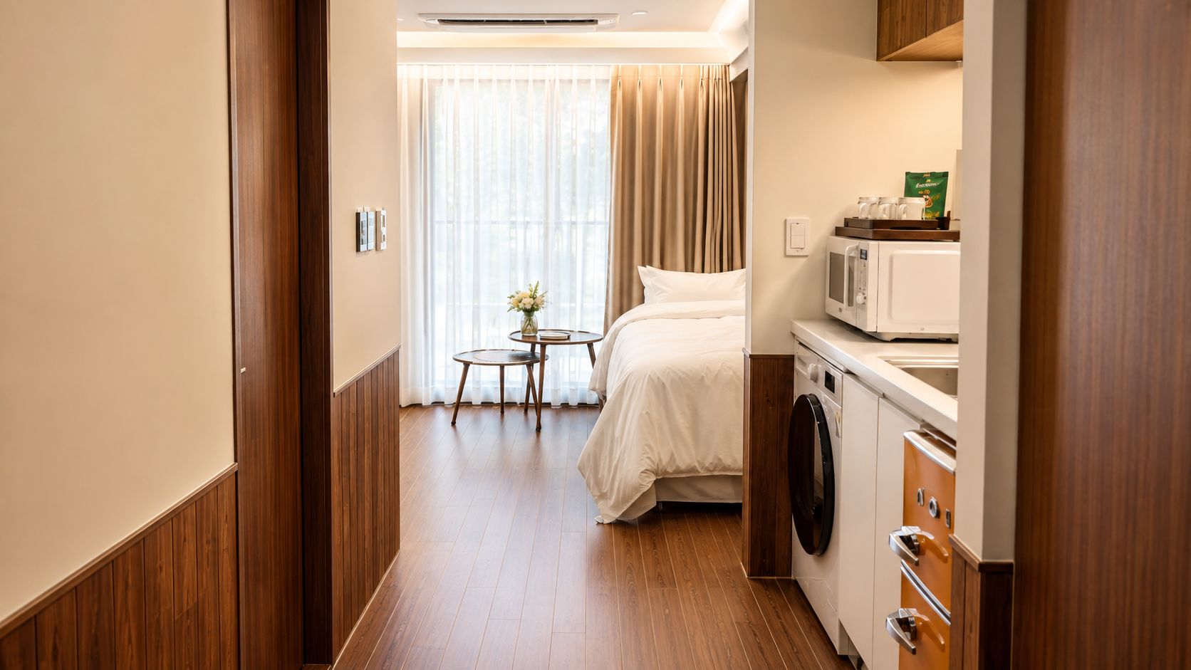 A hotel room with a bed, a washing machine, a microwave, and a small table with a vase of flowers.