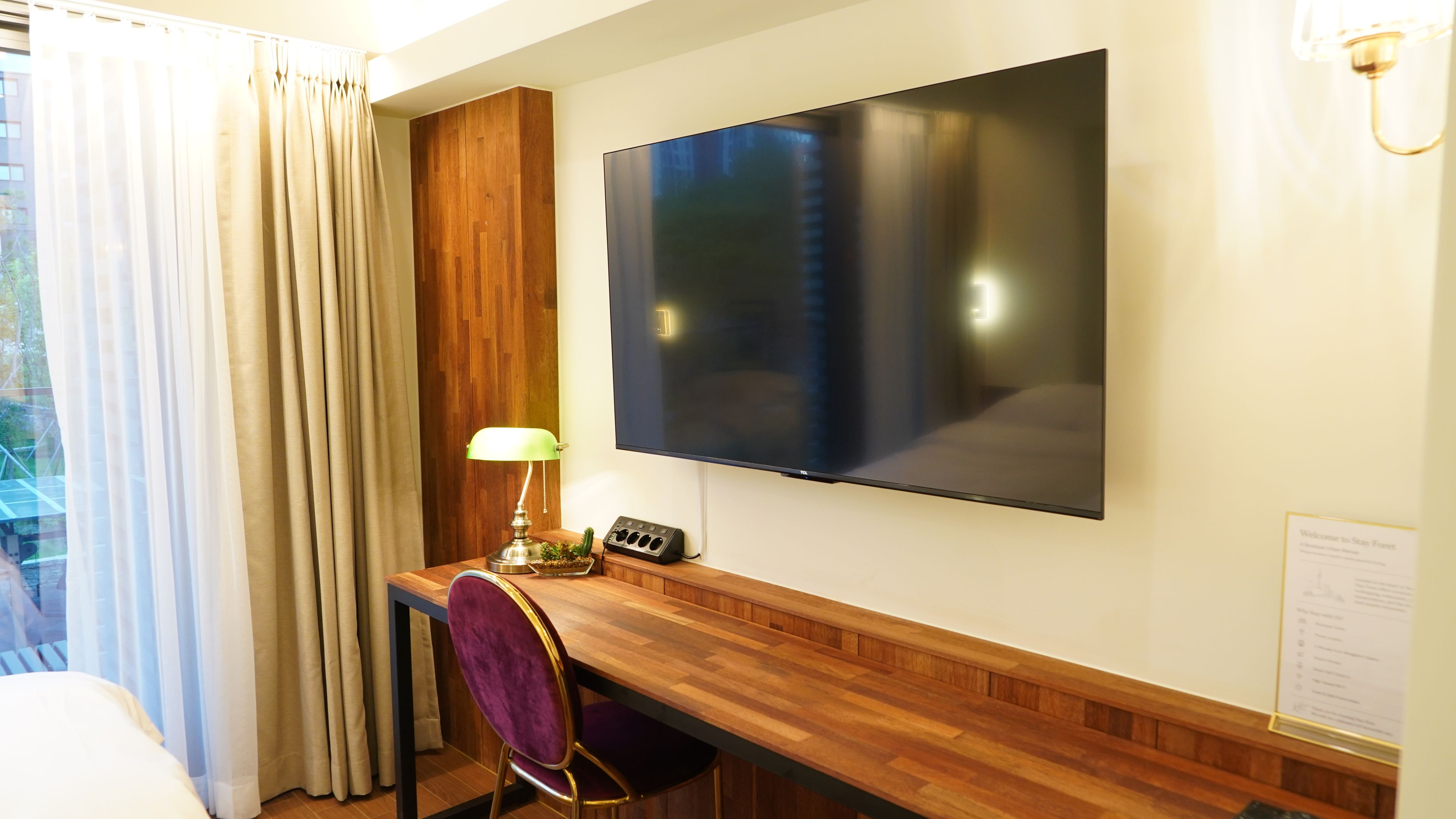 A hotel room with a wooden desk, a flat-screen TV mounted on the wall, a lamp, a chair, and a window with curtains.